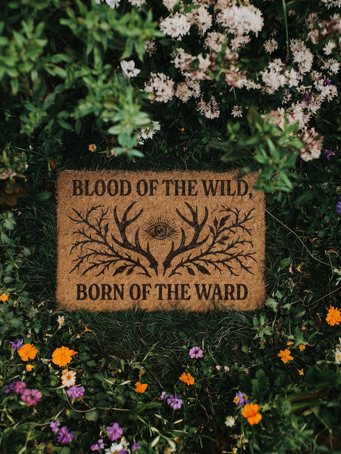 Rectangular coir doormat measuring 24x16 inches featuring black printed text reading “Blood of the Wild, Born of the Ward” with an all-seeing eye symbol and antler-branch illustration. Made from natural coconut fiber with a durable black vinyl backing, this outdoor doormat is weather-resistant and ideal for gothic, witchy, or folklore-inspired entryways.
